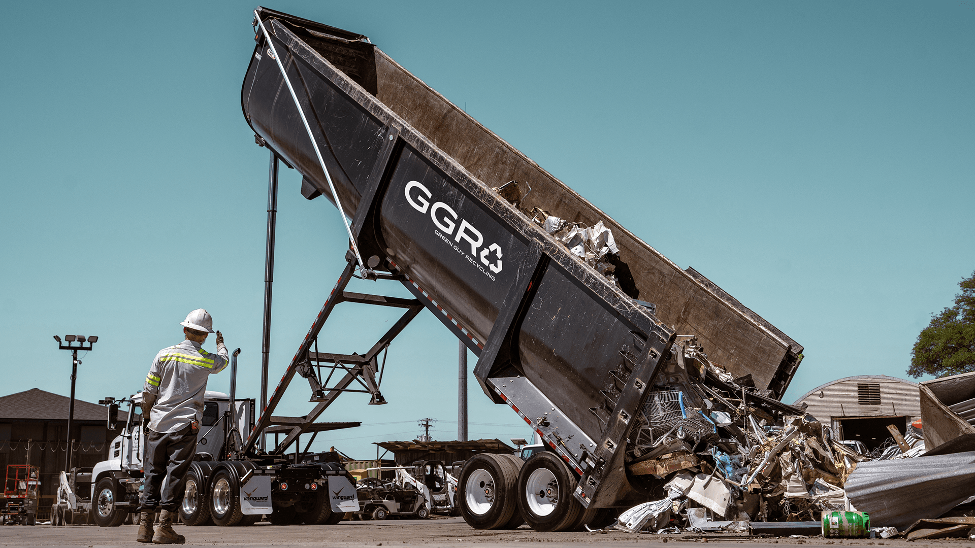 GGR worker directing a roll-off truck as it unloads a large container of scrap metal at a recycling facility, with 'GGR' logo visible on the container.
