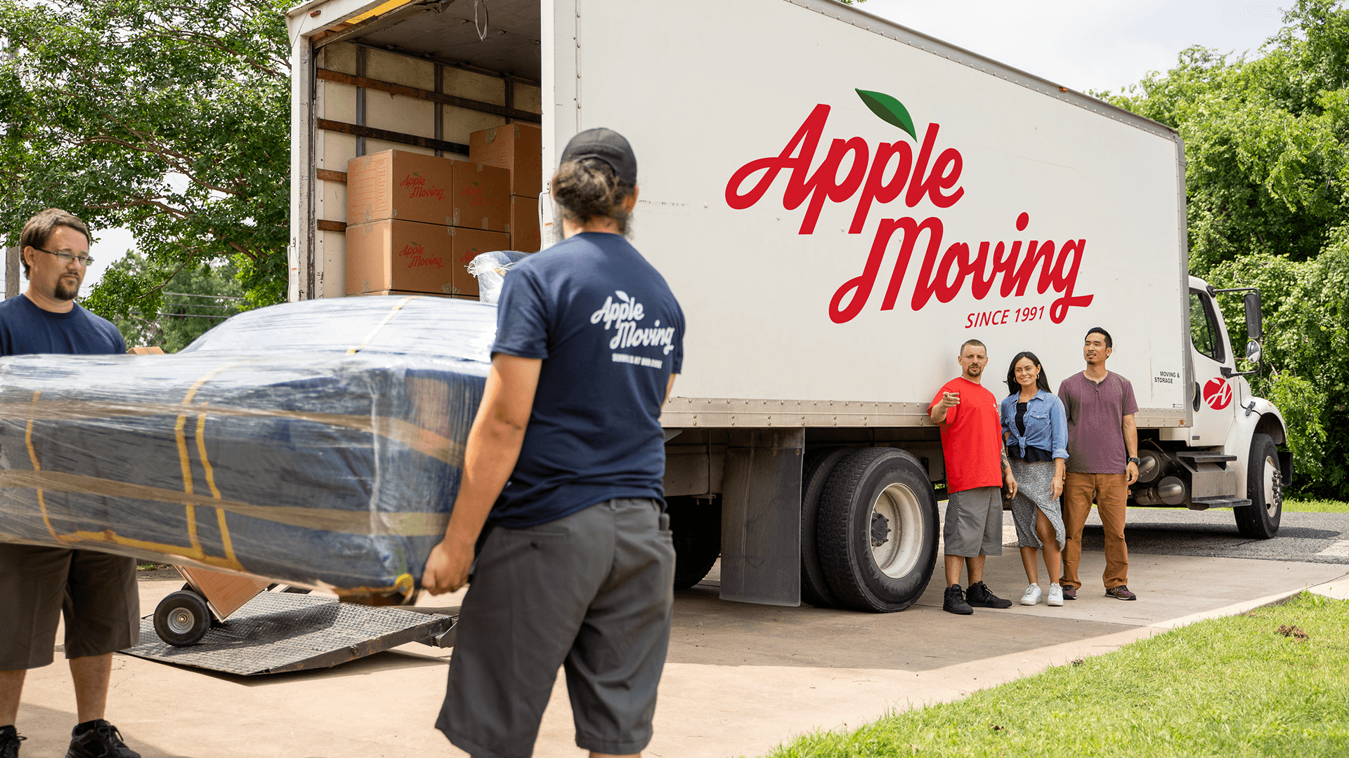 Professional movers from Apple Moving loading a wrapped sofa into a moving truck while a satisfied family looks on outside their home.