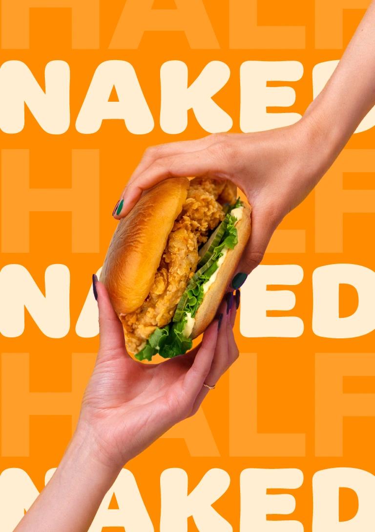 Two hands holding a crispy fried chicken sandwich with lettuce and sauce on a toasted bun against a bright orange background patterned with repeated words.