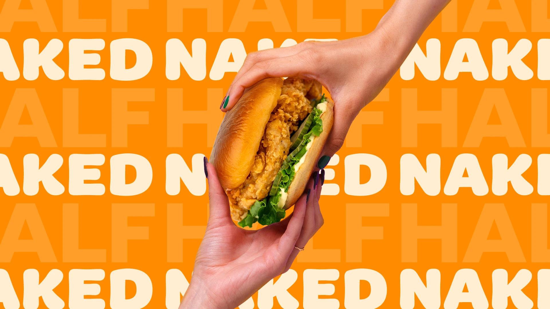 Two hands holding a crispy fried chicken sandwich with lettuce and sauce on a toasted bun against a bright orange background patterned with the repeated words “HALF NAKED” (describing the name of the sandwich).